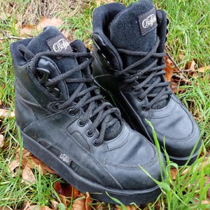 May include: A pair of black Buffalo high-top sneakers. The sneakers have black laces, a padded collar, and the Buffalo logo on the tongue and side. The shoes are sitting on grass and fallen leaves.