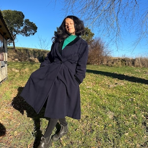 Puede incluir: Una mujer con un abrigo azul marino con cuello de terciopelo negro y un jersey de cuello alto verde. Está de pie en un campo de hierba con una casa al fondo.