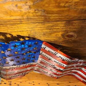 May include: A metal American flag wall art with a distressed look. The flag is waving and the text "A well regulated Militia, being necessary to the security of a free State, the right of the people to keep and bear Arms, shall not be infringed." is inscribed on the red stripes.