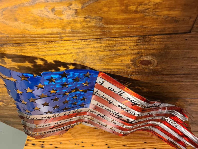 May include: A metal American flag wall art with a distressed look. The flag is waving and the text "A well regulated Militia, being necessary to the security of a free State, the right of the people to keep and bear Arms, shall not be infringed." is inscribed on the red stripes.