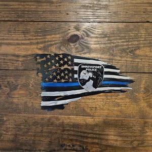 Puede incluir: Un diseño de bandera estadounidense desgastada con una línea azul y la insignia de la policía de Shreveport. La bandera presenta rayas negras y blancas, estrellas y una línea azul, montada sobre un fondo de madera.