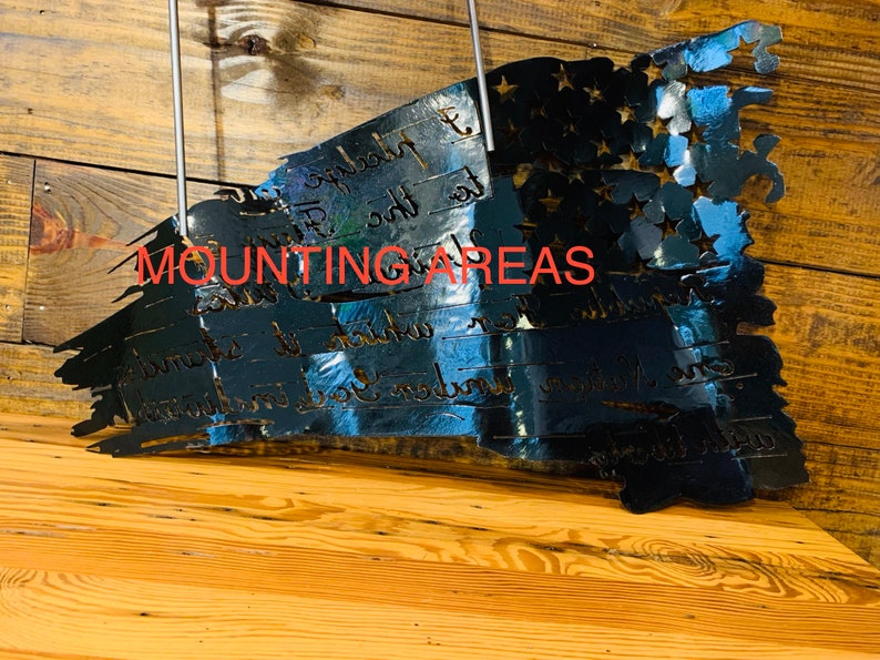 May include: A black metal American flag with a distressed look. The flag is mounted on a wooden wall and has the words "We the People" inscribed on it. The words "Mounting Areas" are written in red text at the top of the image.
