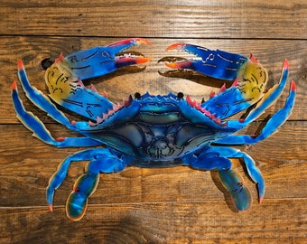 May include: A blue metal crab sculpture with red, yellow, and orange accents. The crab is posed with its claws raised and its legs spread out. The sculpture is mounted on a wooden background.