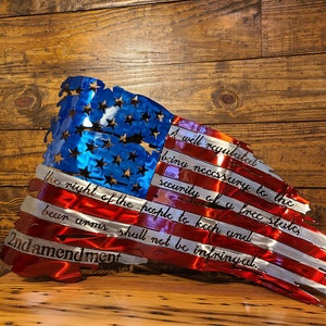 May include: A distressed American flag metal wall art with the 2nd Amendment text: "A well regulated Militia, being necessary to the security of a free State, the right of the people to keep and bear Arms, shall not be infringed."