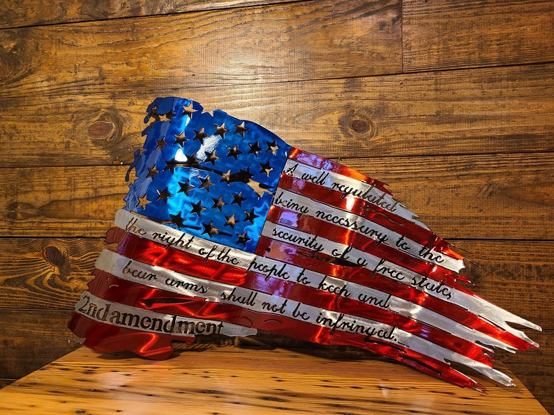 May include: A distressed American flag metal wall art with the 2nd Amendment text: "A well regulated Militia, being necessary to the security of a free State, the right of the people to keep and bear Arms, shall not be infringed."