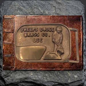May include: A rectangular brass belt buckle featuring a raised relief design of a person pouring liquid from a container. The text PHELPS DODGE BRASS CO. LEE is embossed on the surface. The buckle has a metallic finish with a smooth rectangular area.
