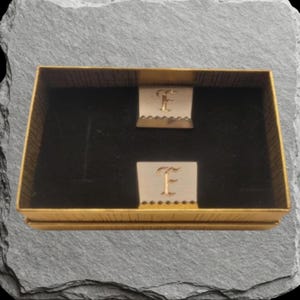 May include: A pair of rectangular metallic cufflinks featuring an engraved letter F in a serif font. The cufflinks are presented in a gold-toned box with a black velvet interior lining, designed to hold the two pieces securely in place.
