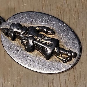 May include: Silver-toned oval pendant with a detailed relief of a figure holding a staff. The pendant has a textured surface and is attached to a metal chain. The figure is in dark tones, with gold accents.