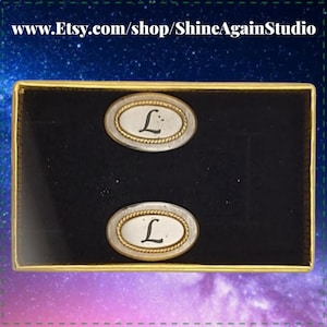 May include: A pair of oval cufflinks with the letter "L" in black script on a silver background, framed by a gold rope design. The cufflinks are set in a black velvet box with a gold border.