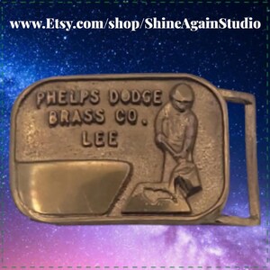 May include: A vintage brass belt buckle with the text "PHELPS DODGE BRASS CO. LEE". The buckle features a relief of a person working with a hammer and anvil. The buckle is rectangular with a rounded top and a rectangular loop.