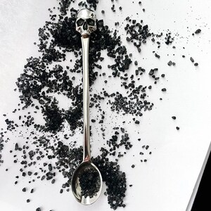 May include: A silver skull-shaped spoon with a black salt spoon on a white surface.