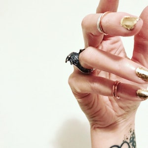 May include: A hand wearing a silver ring and a large, detailed, black ring with a textured surface. The hand is also wearing a thin, copper band on the pinky finger. The nails are painted with gold glitter.