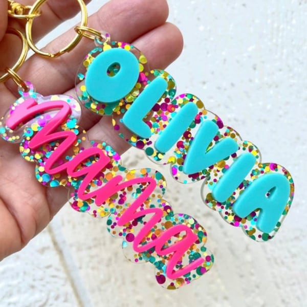 May include: Two clear acrylic keychains with the names "Olivia" and "mama" spelled out in colorful glitter. The letters are in a bubble font and the glitter is in shades of pink, blue, green, gold, and purple.