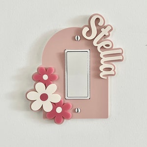 May include: A light switch cover with a pink arch design and the name "Stella" in white lettering. The cover has three pink and white flowers on the left side.