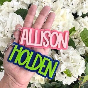 May include: Two personalized keychains with the names "Allison" and "Holden" in bold, colorful letters. The keychain with "Allison" is pink with white lettering, and the keychain with "Holden" is blue with green lettering.