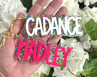 May include: Two colorful resin keychains with the names "Cadance" and "Hadley" spelled out in white and pink letters, respectively. The keychains are filled with colorful confetti.