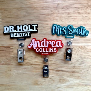 May include: Three custom name badge reels with metal clips. One black with white text reading DR. HOLT DENTIST, one red with pink text reading Andrea COLLINS, and one teal with dark teal script text reading Mrs. Smith.