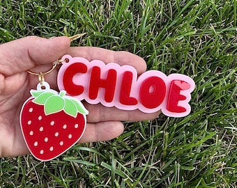May include: A strawberry-shaped keychain with a red and white design, featuring a green leaf and a gold keyring. The keychain also includes the name "CHLOE" in red letters on a pink background.