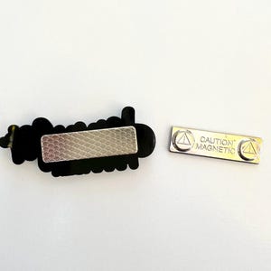 Puede incluir: Una placa de identificaci&oacute;n negra con la palabra "hello" en fuente cursiva. La placa de identificaci&oacute;n tiene un respaldo magn&eacute;tico plateado. Tambi&eacute;n se incluye una pieza magn&eacute;tica plateada separada con el texto "CAUTION MAGNETIC".
