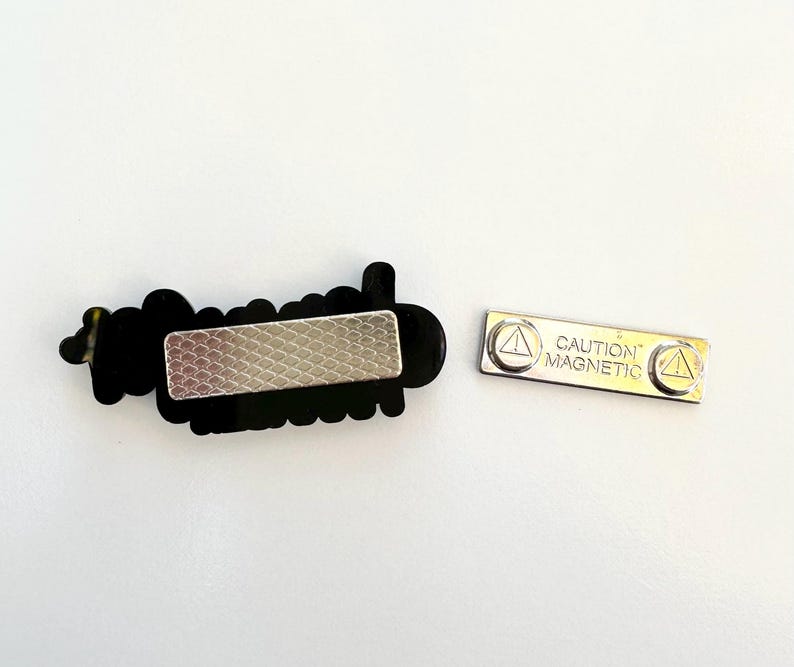 Puede incluir: Una placa de identificaci&oacute;n negra con la palabra "hello" en fuente cursiva. La placa de identificaci&oacute;n tiene un respaldo magn&eacute;tico plateado. Tambi&eacute;n se incluye una pieza magn&eacute;tica plateada separada con el texto "CAUTION MAGNETIC".
