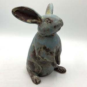Op de afbeelding: Een keramieken konijnfiguur in zittende houding, met een verweerde blauw-bruine afwerking. Het konijn heeft lange oren en een gedetailleerd gezicht, voor interieurdecoratie.