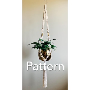 Può includere: Un portavasi in macramé bianco con un vaso dorato e una pianta verde. Il portavasi ha un fiocco in basso e il testo "Pattern" è visibile sotto il vaso.
