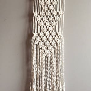 May include: Off-white macrame wall hanging. The handmade piece features a geometric pattern with knotted details and long fringe. It is suspended from a natural wooden dowel, adding a touch of bohemian decor to any space.