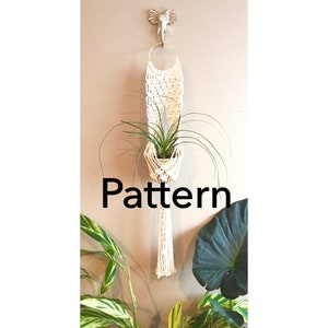 May include: A white macrame plant hanger with a small potted plant hanging from it. The hanger is attached to a wall with a gold ring. The word "Pattern" is written in black text below the hanger.