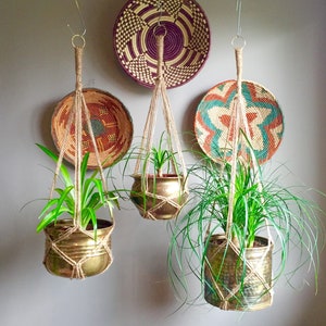 May include: Three hanging macrame plant hangers with brass pots and green plants. The hangers are made of natural twine and are hanging from hooks on a white wall. There are three woven baskets on the wall behind the hangers.