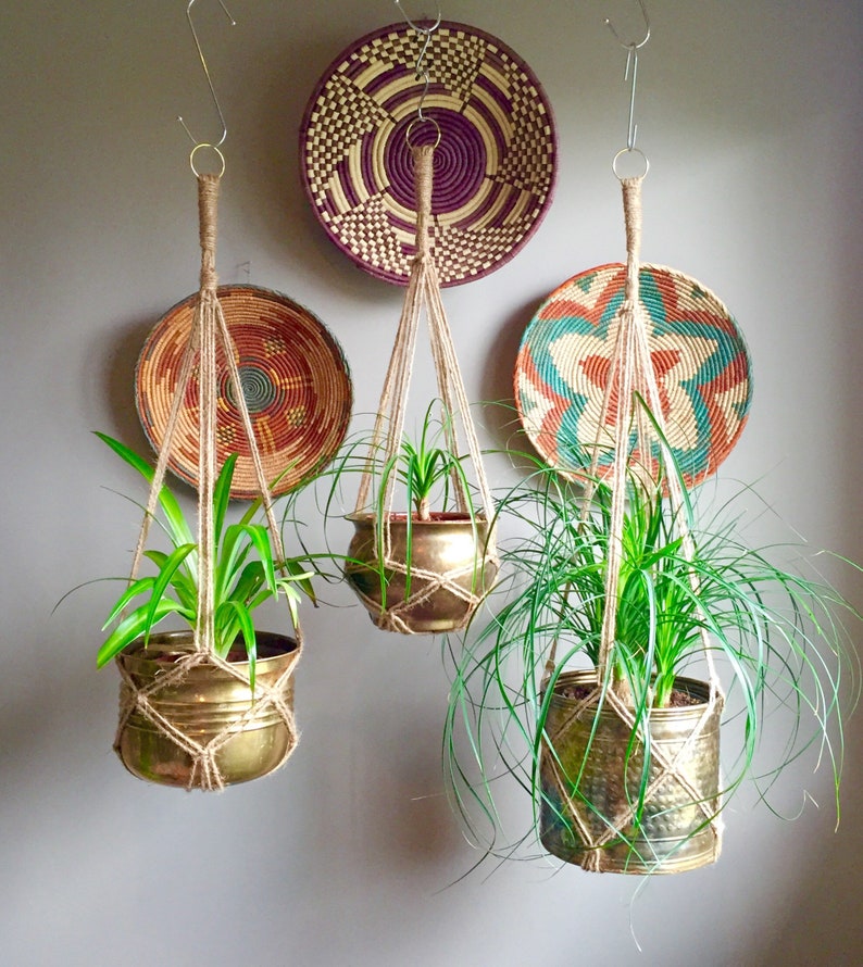 May include: Three hanging macrame plant hangers with brass pots and green plants. The hangers are made of natural twine and are hanging from hooks on a white wall. There are three woven baskets on the wall behind the hangers.