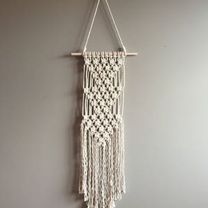 May include: A cream-colored macrame wall hanging. The handmade piece features a geometric pattern with a wooden dowel at the top and long fringe at the bottom. It is suspended from a decorative metal hook on a gray wall.