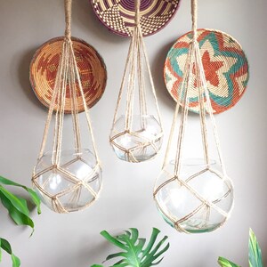 May include: Three macrame plant hangers with glass globes hanging from hooks on a wall. The hangers are made of natural twine and the globes are clear glass. There are three woven baskets on the wall behind the hangers.