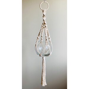May include: A white macrame plant hanger with a glass sphere for a planter. The hanger has a tassel at the bottom.
