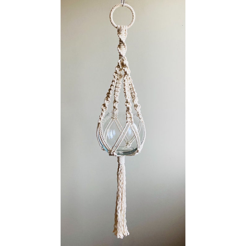 May include: A white macrame plant hanger with a glass sphere for a planter. The hanger has a tassel at the bottom.
