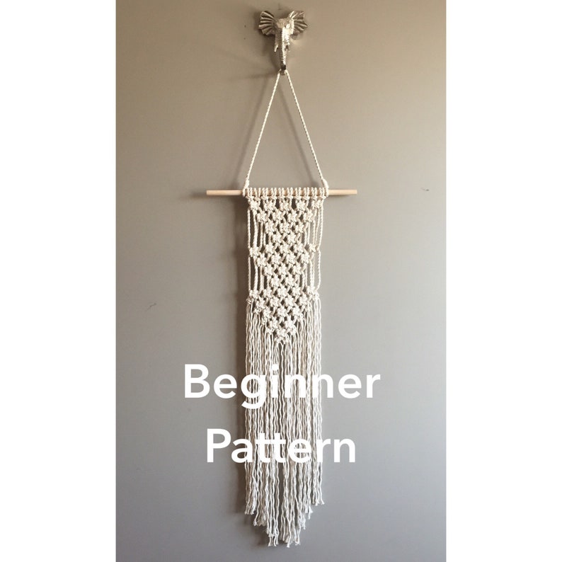 May include: A macrame wall hanging with a wooden dowel and fringe. The off-white piece is suspended from a decorative elephant hook. The text "Beginner Pattern" is displayed on the wall hanging.