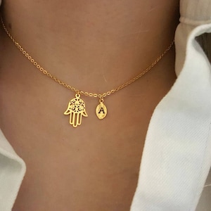 Könnte beinhalten: Eine goldene Kettenkette mit zwei Anhängern: einer Hamsa-Hand und einem Buchstaben 'A'.