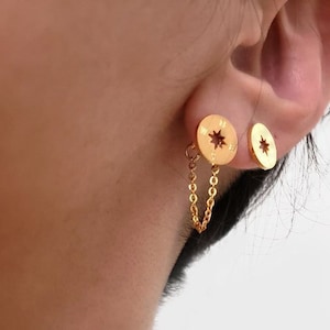 May include: Gold compass earrings with a dangling chain. The earrings are in the shape of a compass with a star in the center. The chain is attached to the bottom of the compass.