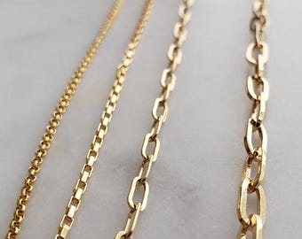 May include: Close-up of three gold necklaces with different chain styles. The chains include a delicate beaded chain, a box chain, and a larger oval link chain. The necklaces are displayed on a light-colored surface.