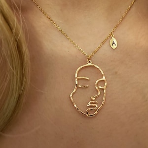 Peut inclure: Un collier en or avec une chaîne délicate et un pendentif représentant un dessin minimaliste d'un visage. Un petit pendentif avec la lettre 'A' pend de la chaîne.