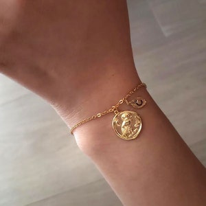Puede incluir: Una pulsera de oro con un dije de mono y un dije de ojo turco. La pulsera está en la muñeca de una persona.