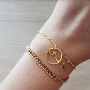 May include: Two gold bracelets on a wrist. One bracelet is a delicate chain with a wave charm. The other bracelet is a thicker curb chain.