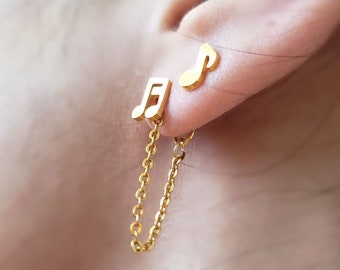 May include: A pair of gold-colored musical note earrings. One earring is a single note stud, and the other is a double note stud connected by a delicate chain. The earrings are displayed on an ear, showcasing their design and color.