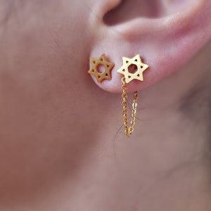 Puede incluir: Pendientes dorados en forma de estrella. Un pendiente es un perno y el otro está conectado por una delicada cadena. Los pendientes tienen un diseño simple y elegante. Los pendientes se llevan en la oreja.