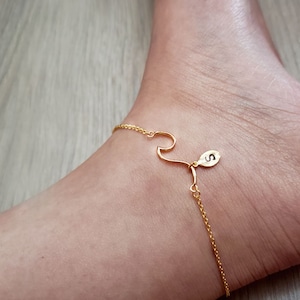 May include: Gold ankle bracelet with a wave charm and a small charm with the letter 'S'.