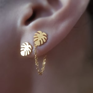 以下が含まれることがあります： ゴールドトーンのイヤリングは、つながったモンステラの葉のチャームが2つ付いています。葉は繊細なゴールドチェーンでつながっています。