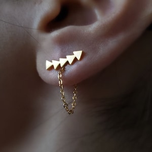 May include: Gold triangle shaped earring with a chain dangling from the bottom. The earring is worn in the earlobe.