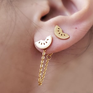 以下が含まれることがあります： 2つのパーツを繋ぐチェーンが付いた、金色のスイカの輪切り形のイヤリング。イヤリングは人の耳につけられています。