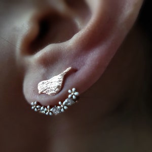 Puede incluir: Un par de pendientes de oro con un pequeño diseño de pájaro y una fila de pequeñas flores blancas.