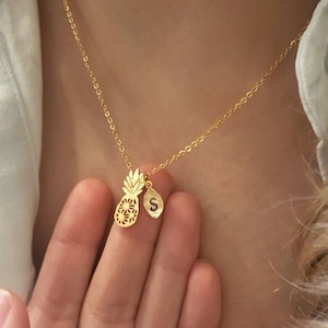 Peut inclure: Un collier en chaîne dorée avec un pendentif ananas et un petit pendentif avec la lettre 'S' qui pend.