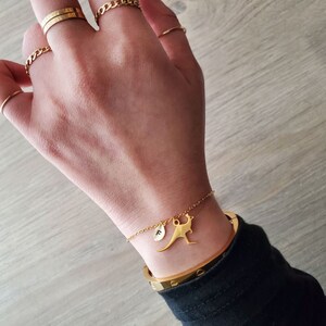 Puede incluir: Un brazalete de cadena dorada con un pequeño dije de canguro dorado y un dije de inicial dorada. El brazalete está en la muñeca de una persona.